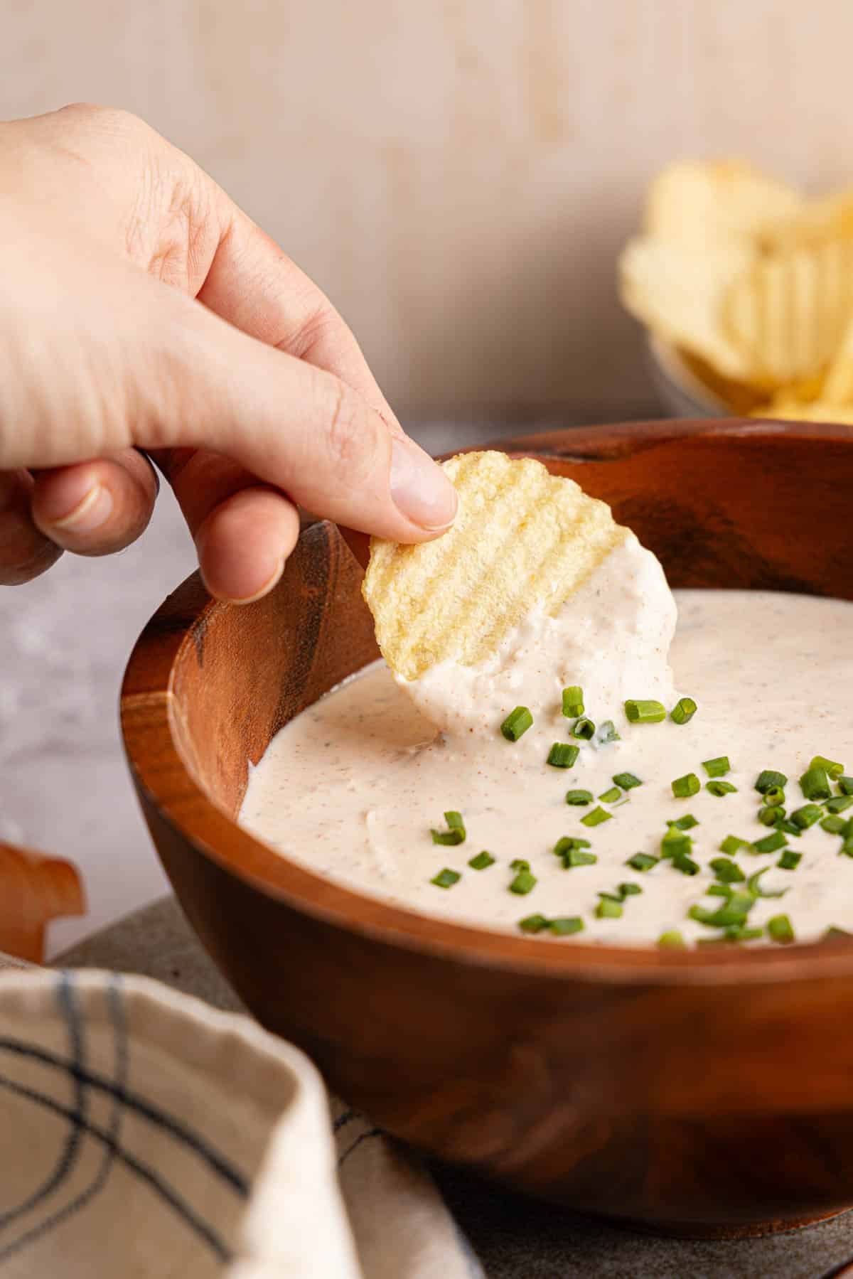 A hand dipping a chip into the greek yogurt french onion dip which is served in a wooden bowl.