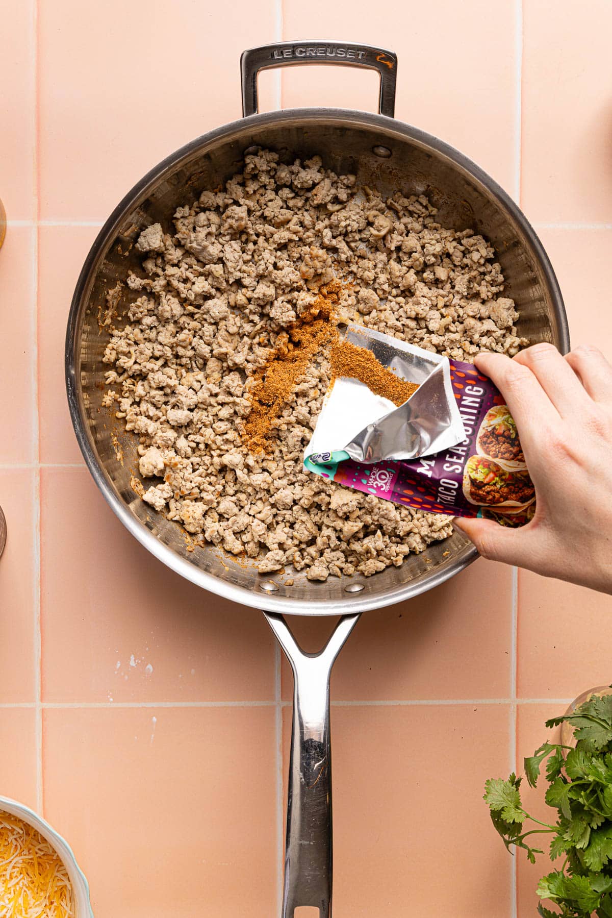 A hand pours the taco seasonings over the cooked ground turkey in a skillet.