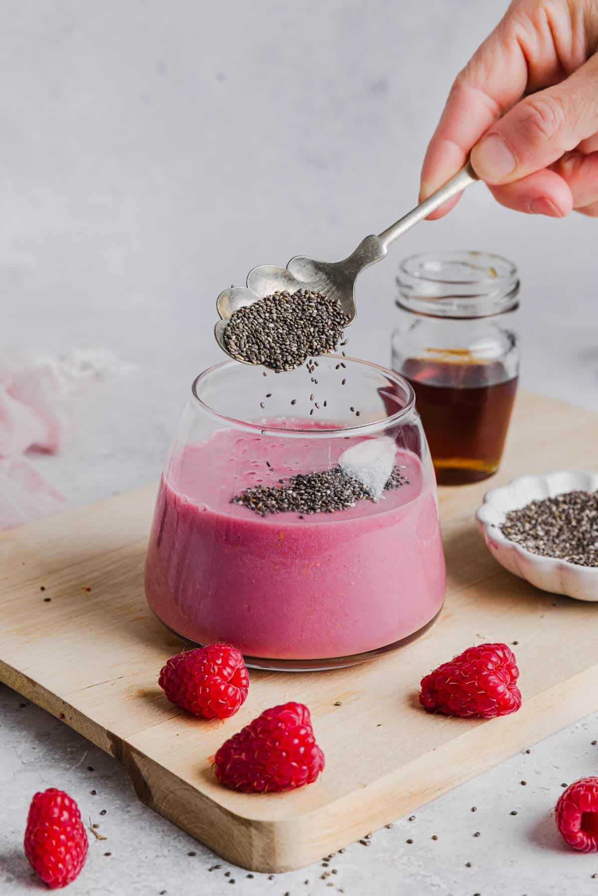 A spoon is pouring chia seeds into a glass filled with the blended raspberry mixture.