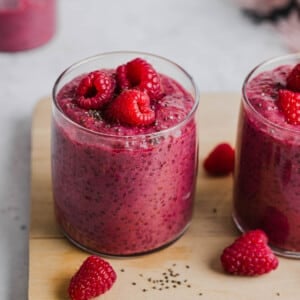 A glass filled with raspberry chia pudding and fresh raspberries on too sitting on top of a wooden cutting board.