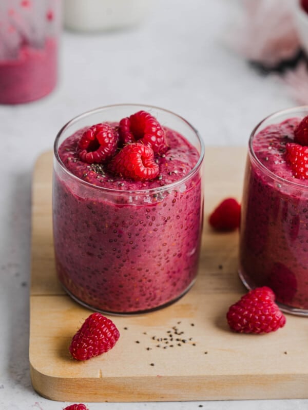 A glass filled with raspberry chia pudding and fresh raspberries on too sitting on top of a wooden cutting board.