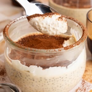 A spoon with a spoonful of tiramisu pudding over a bowl filled with the pudding.