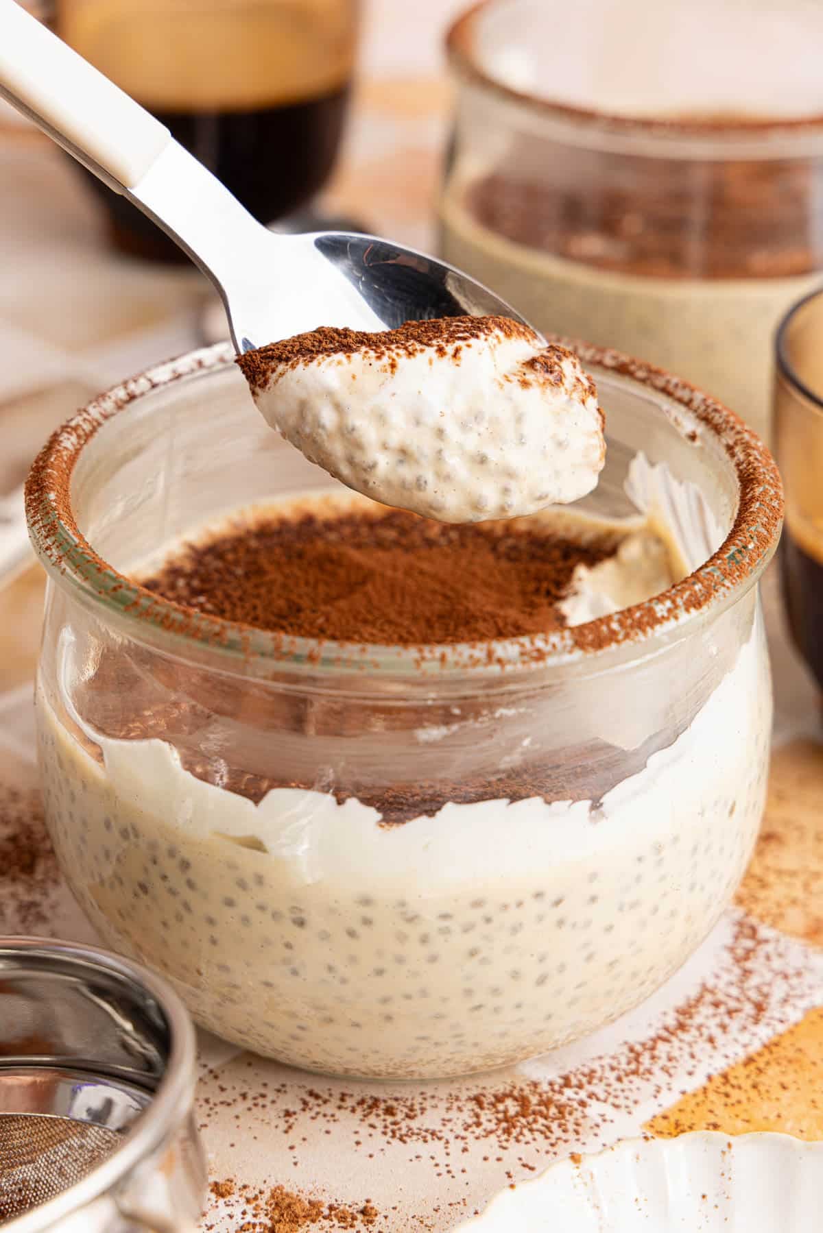 A spoon with a spoonful of tiramisu pudding over a bowl filled with the pudding.
