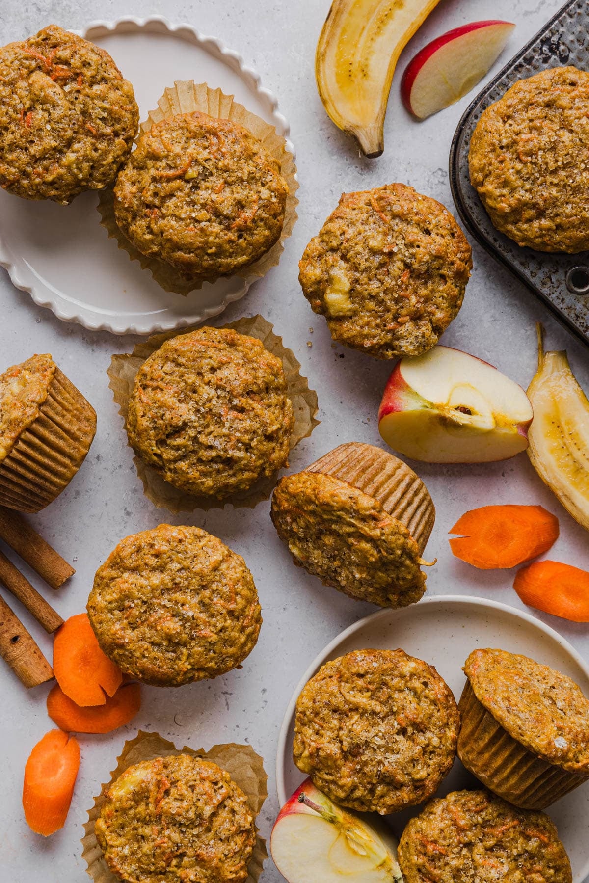 The baked apple banana carrot muffins on a countertop with banana apple slices and carrot slices.