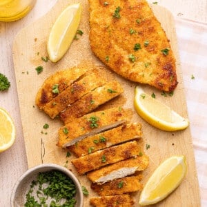 Two chicken cutlets on a wooden cutting board. Once is sliced. There are lemon wedges off to the side.