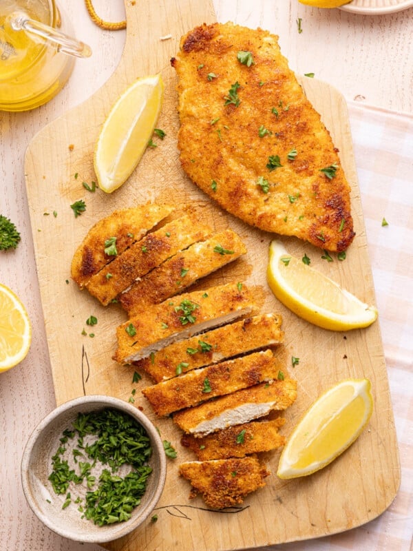 Two chicken cutlets on a wooden cutting board. Once is sliced. There are lemon wedges off to the side.