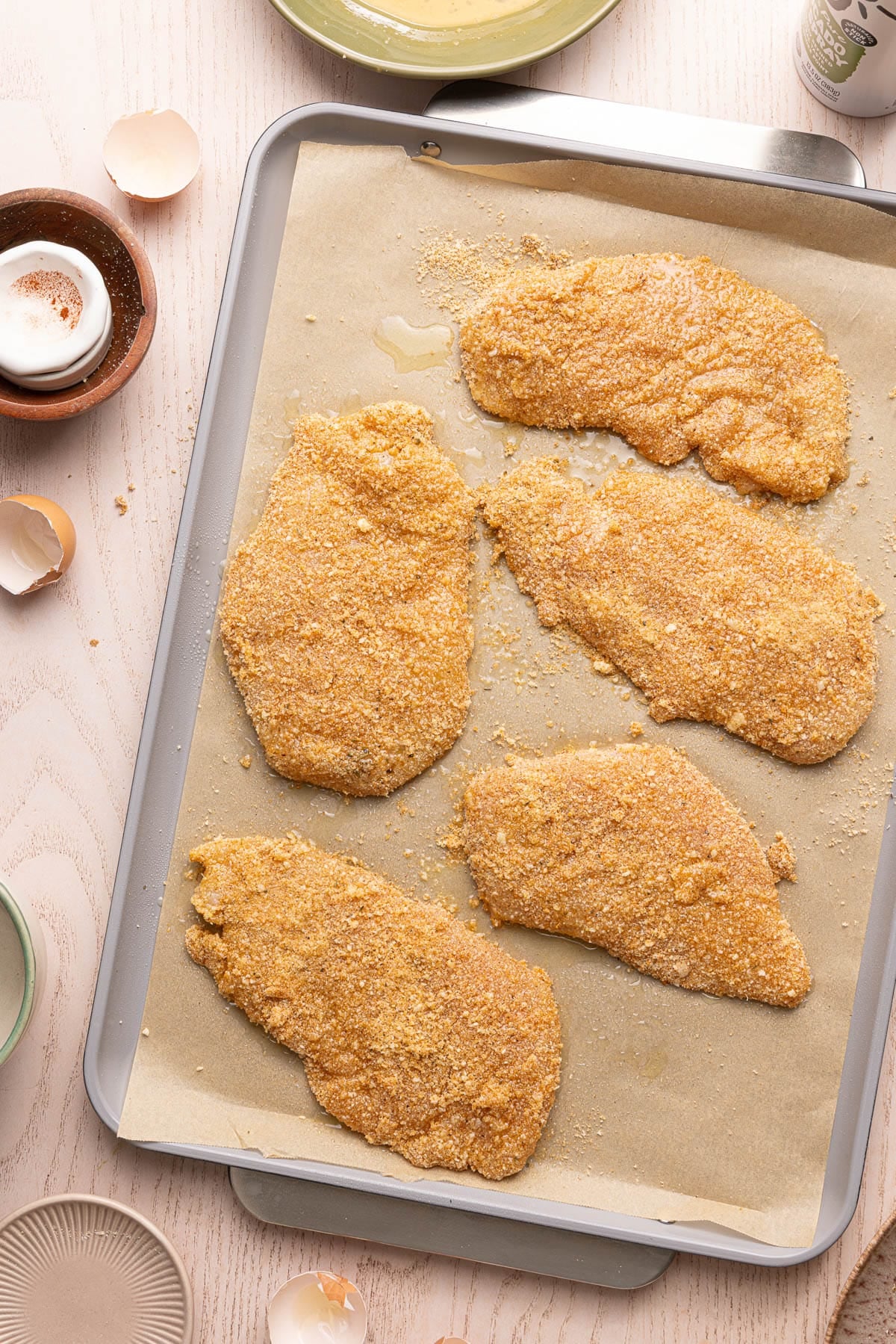 The five chicken cutlets coated in the breading with a splash of oil on top of each on a baking sheet lined with parchment paper.