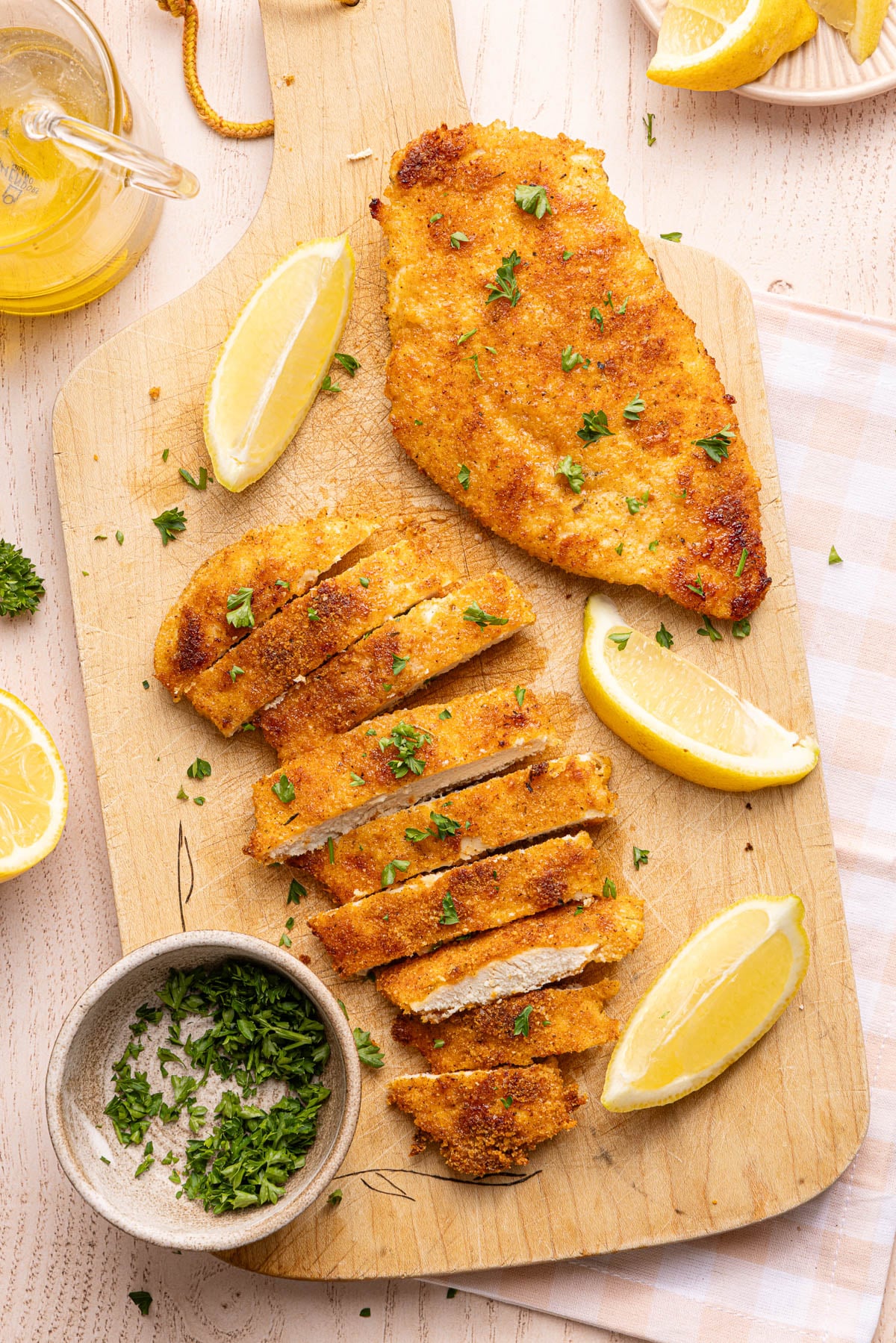 Two chicken cutlets on a wooden cutting board. Once is sliced. There are lemon wedges off to the side.