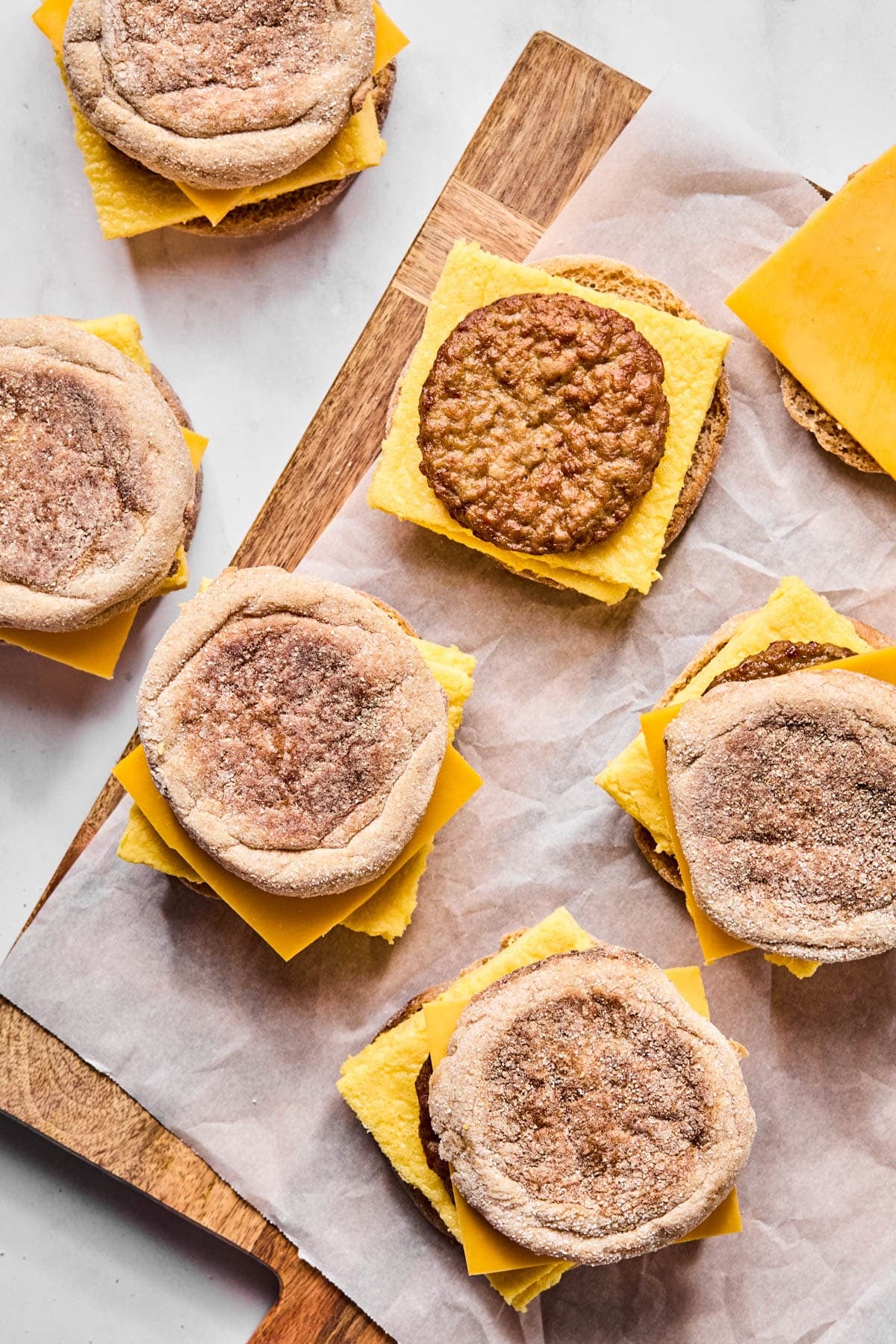 Six breakfast sandwiches sit on a piece of parchment paper on top of a wooden cutting board when assembling.