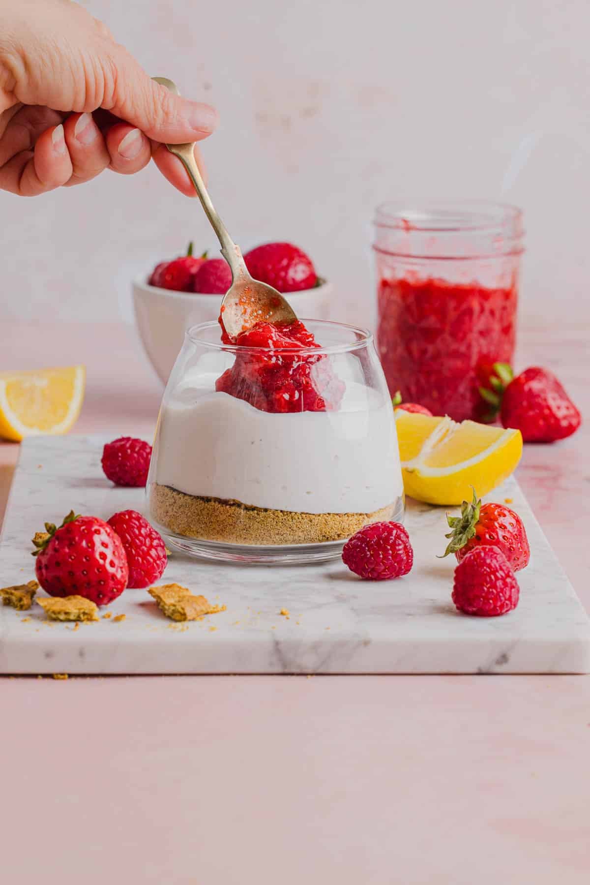 The crushed graham crackers, cheesecake filling, and strawberry topping layered in a glass jar on a white serving platter.