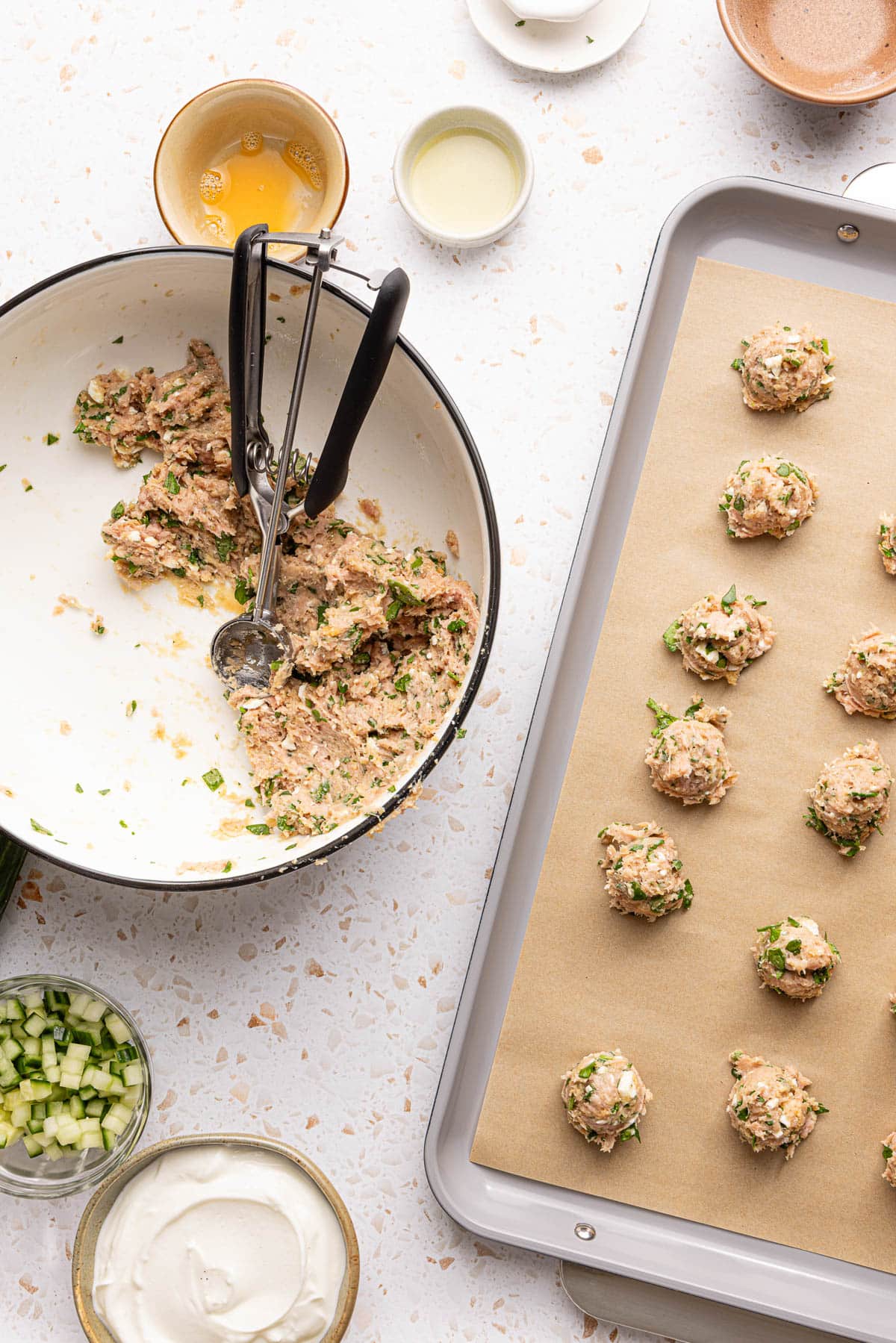 A cookie scoop rests in a bowl with the meatball batter, then a baking sheet with half of the meatballs is to the right of the bowl on a countertop.