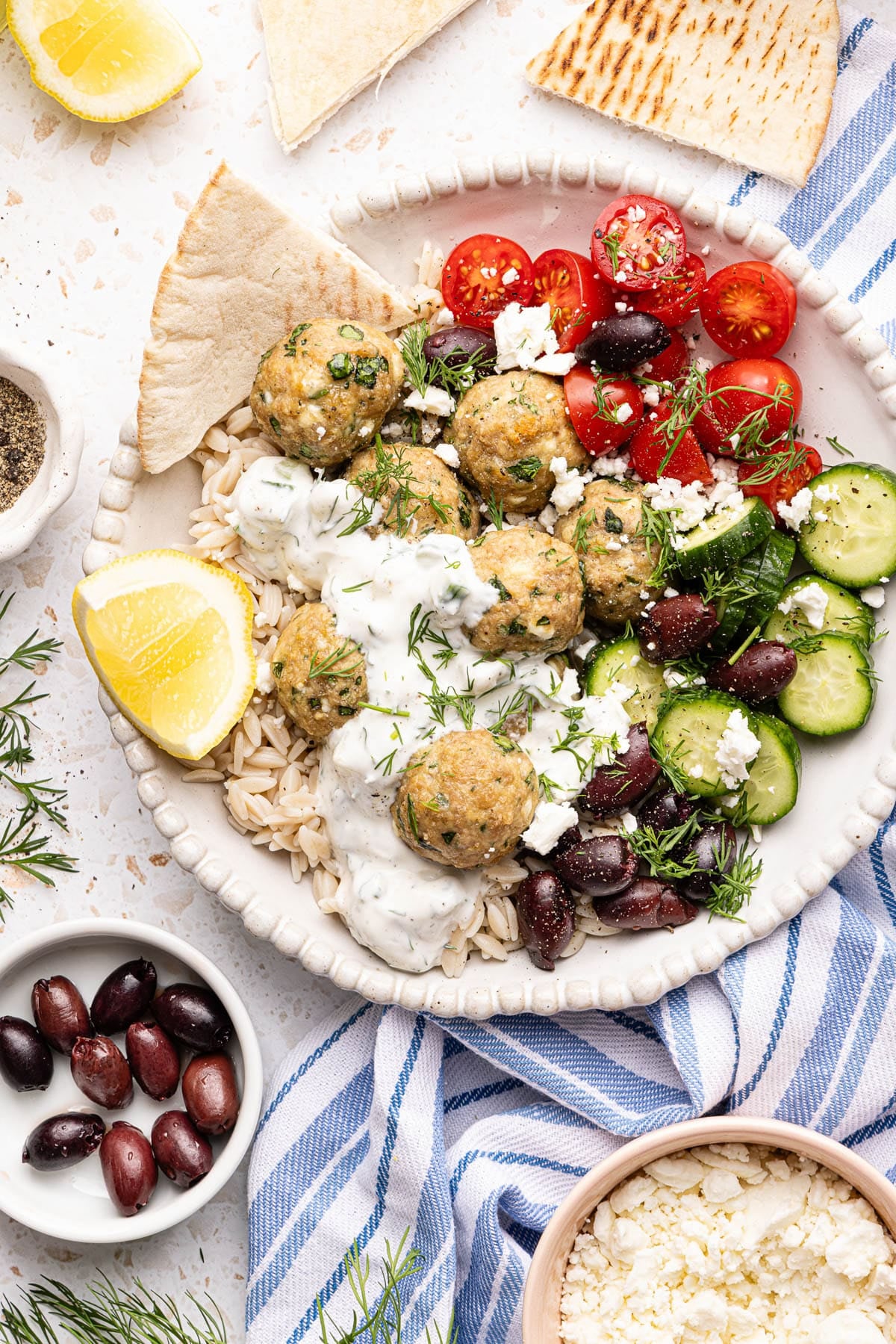 The turkey feta meatballs in a bowl with tomatoes, olives, cucumbers, feta and the yogurt sauce in a bowl with a side of pita and fresh lemon slice.