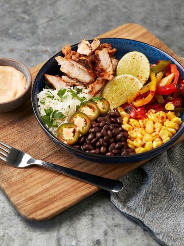 A colorful chicken burrito bowl with black beans, rice, corn salsa, fajita peppers, and jalapeรฑos served with a side of dressing on a wooden board.
