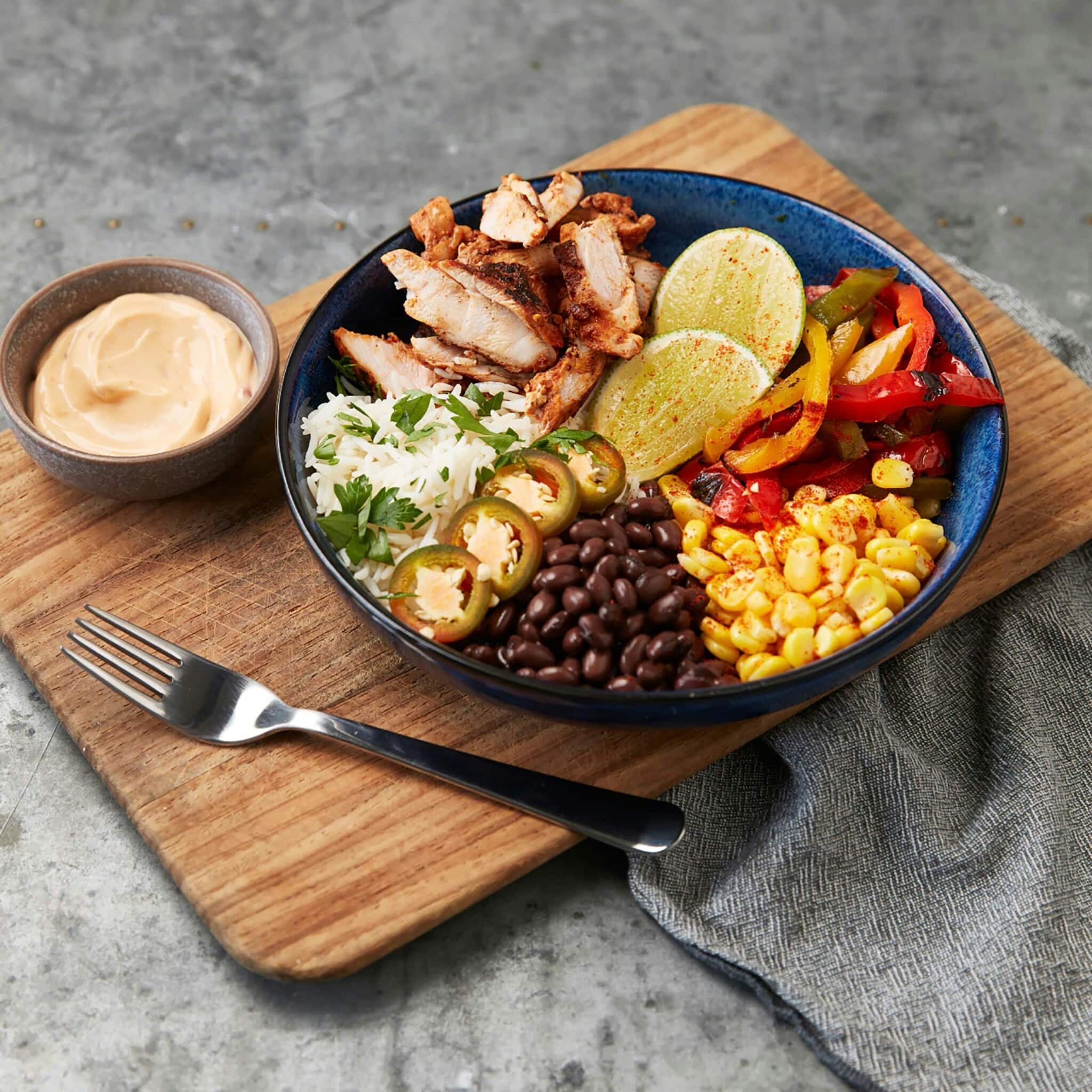 A colorful chicken burrito bowl with black beans, rice, corn salsa, fajita peppers, and jalapeños served with a side of dressing on a wooden board.