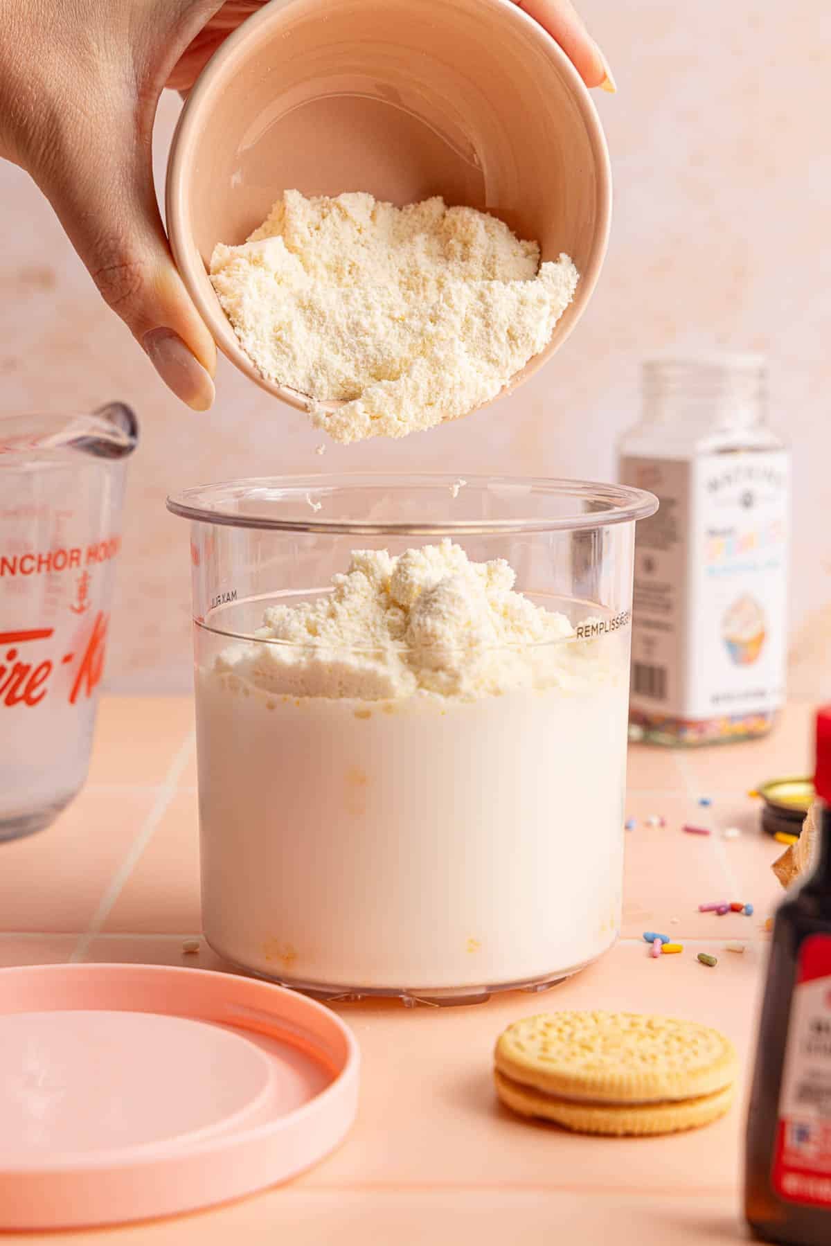 A hand pours the protein powder into the creami pint container with the pudding mix, milk, almond and butter extract before whisking.
