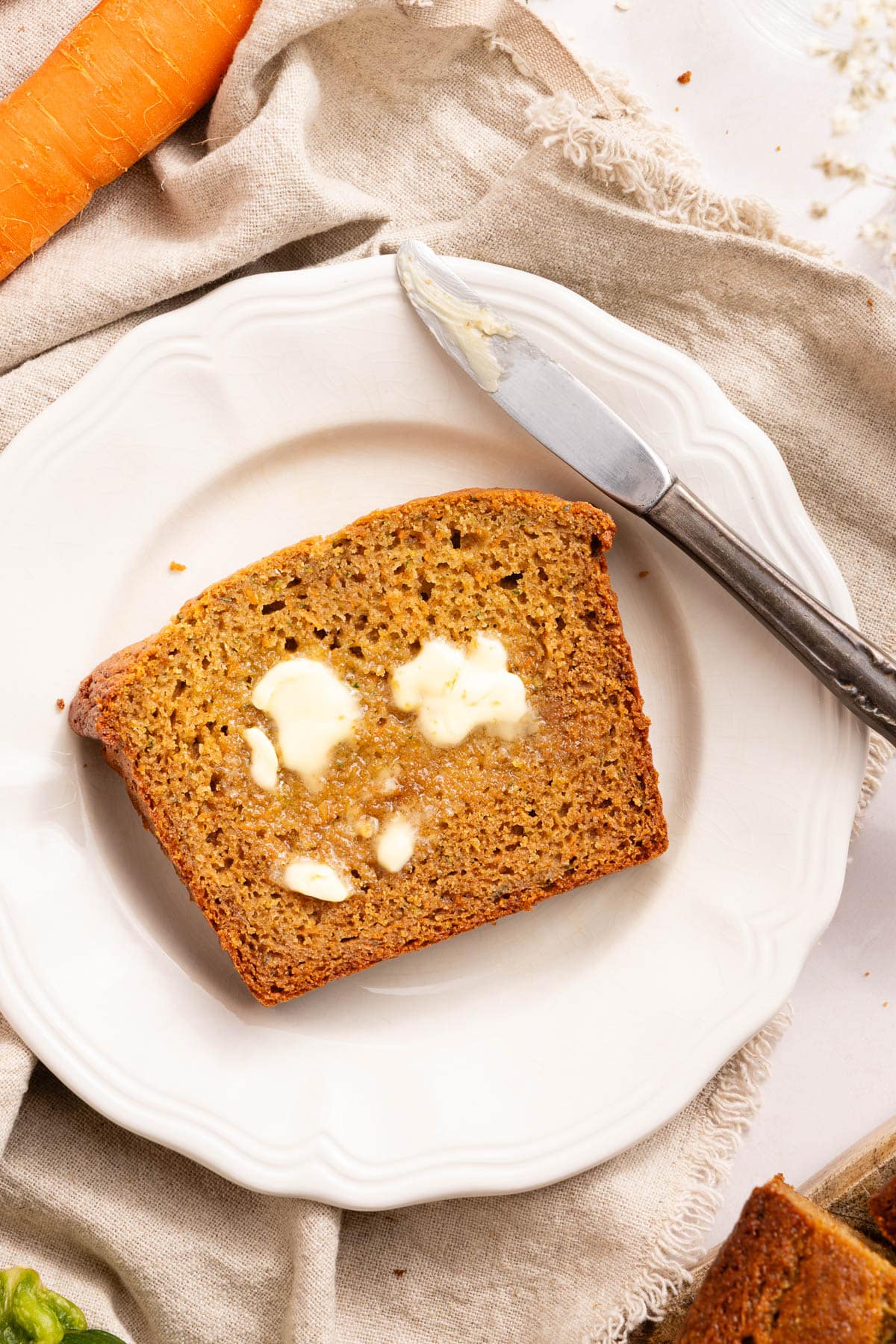 A slice of zucchini and carrot bread on a white plate with a few pats of butter on top.