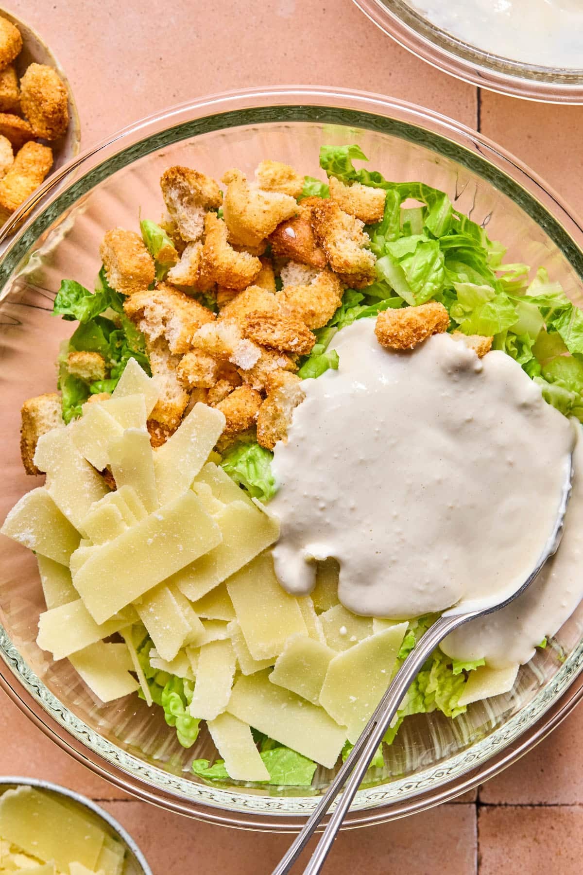 The lettuce, parmesan cheese, and crushed croutons in a large glass bowl with the dressing poured on top.