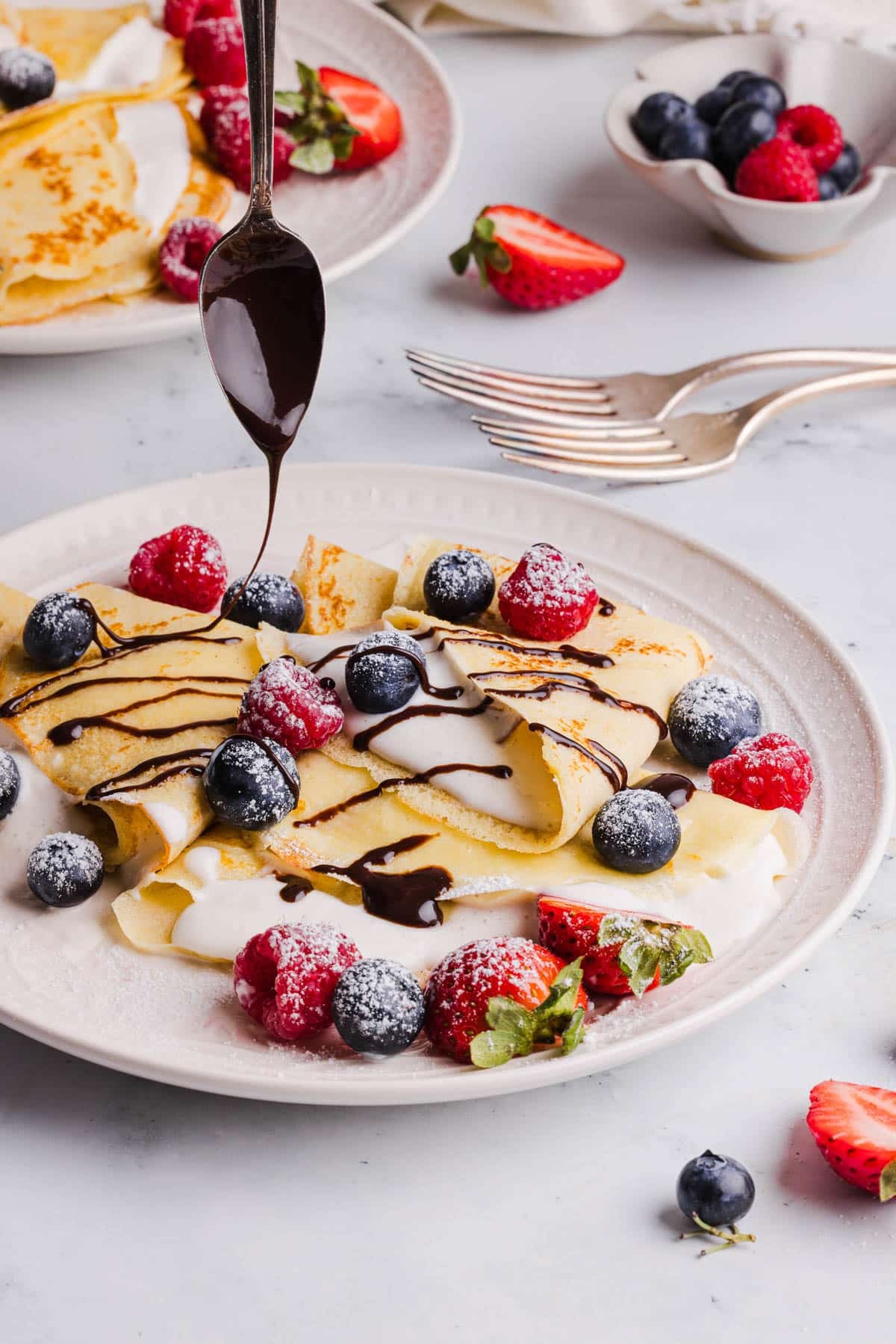 A spoon drizzles chocolate syrup over the crepes on a white plate.
