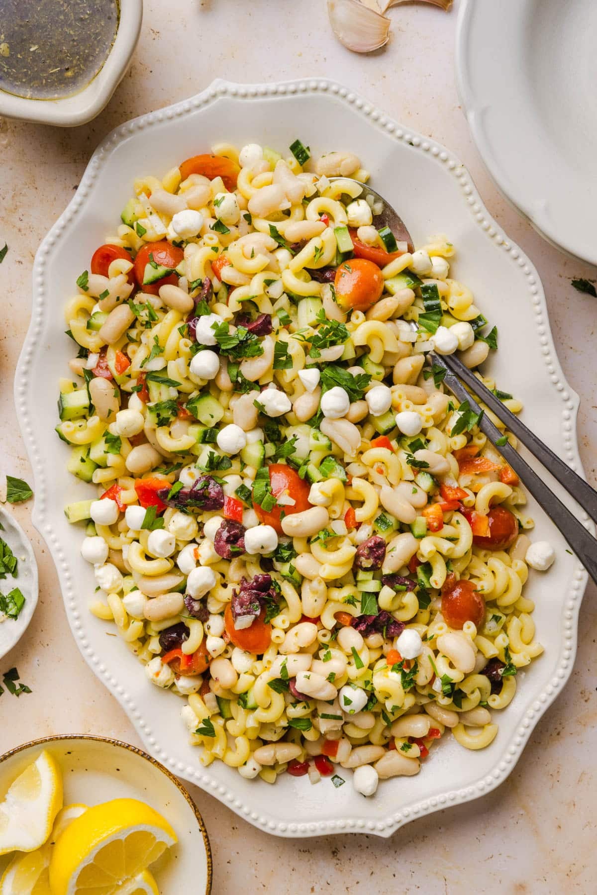 Pasta bean salad on a white oval serving platte with two serving utensils sitting on a countertop.