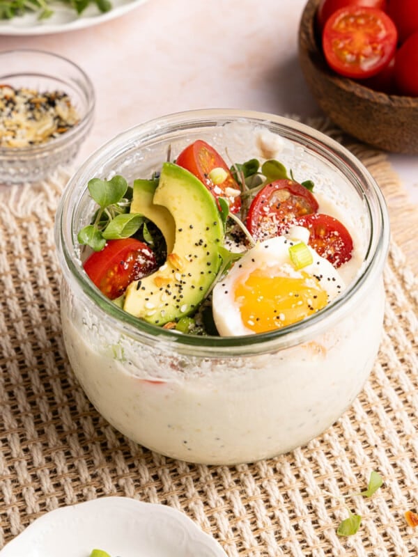 Savory overnight oats in a glass container with the savory toppings on top including sliced tomatoes, avocado, microgreens, and fried egg.