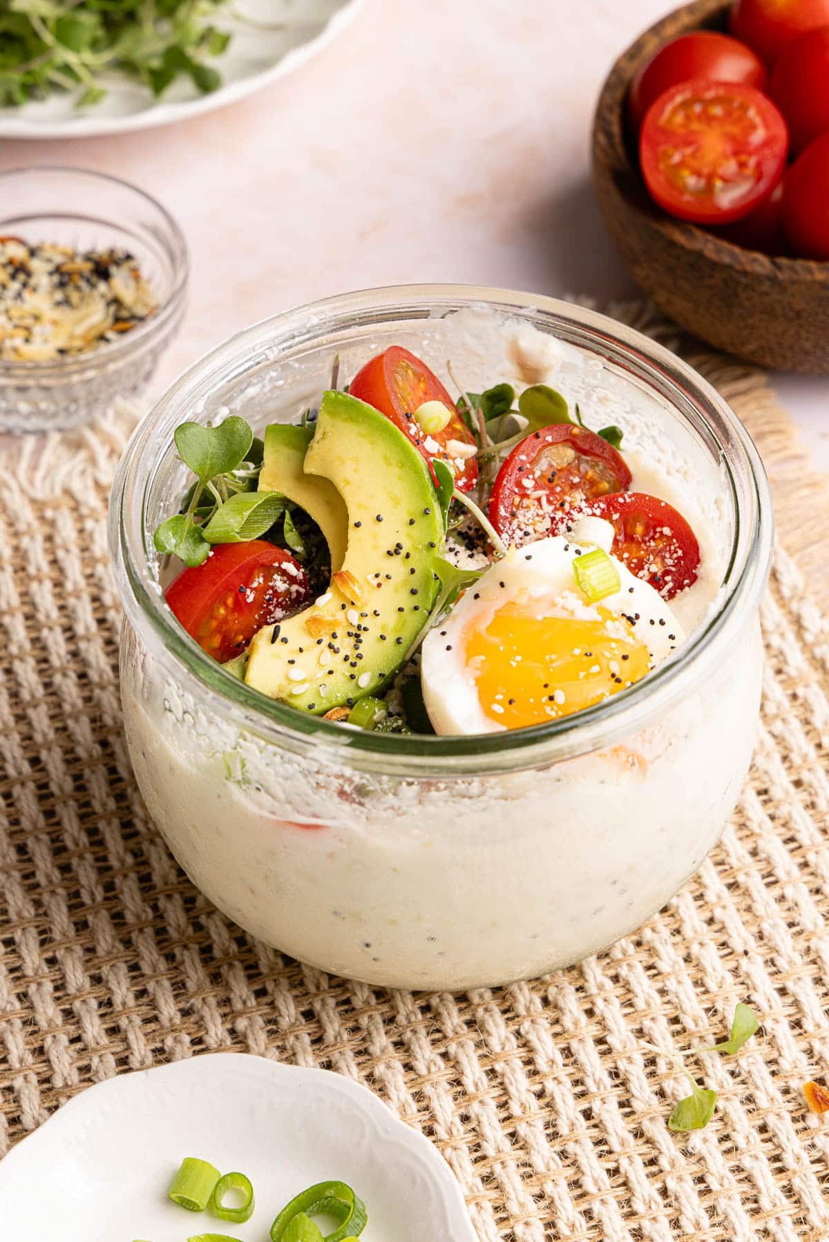 Savory overnight oats in a glass container with the savory toppings on top including sliced tomatoes, avocado, microgreens, and fried egg.