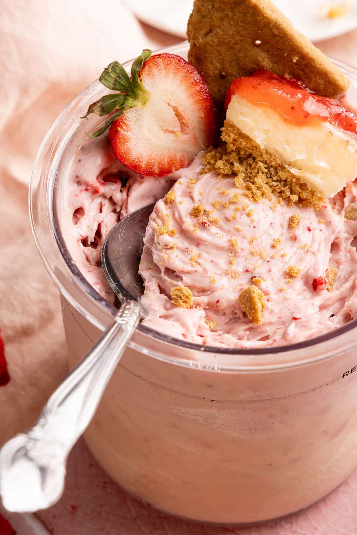 A spoon sits in a container of Ninja Creami Strawberry Cheesecake Ice Cream garnished with a sliced strawberry, graham cracker, and piece of cheesecake. 