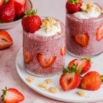 Two jars of strawberry chia pudding garnished with greek yogurt, graham cracker crumbs and fresh strawberries sitting on a white plate on a countertop.
