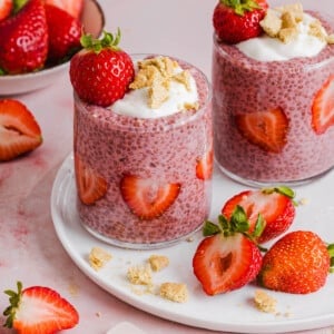 Two jars of strawberry chia pudding garnished with greek yogurt, graham cracker crumbs and fresh strawberries sitting on a white plate on a countertop.
