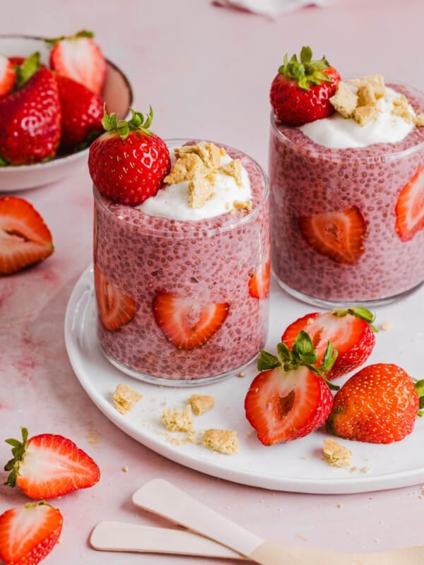 Two jars of strawberry chia pudding garnished with greek yogurt, graham cracker crumbs and fresh strawberries sitting on a white plate on a countertop.
