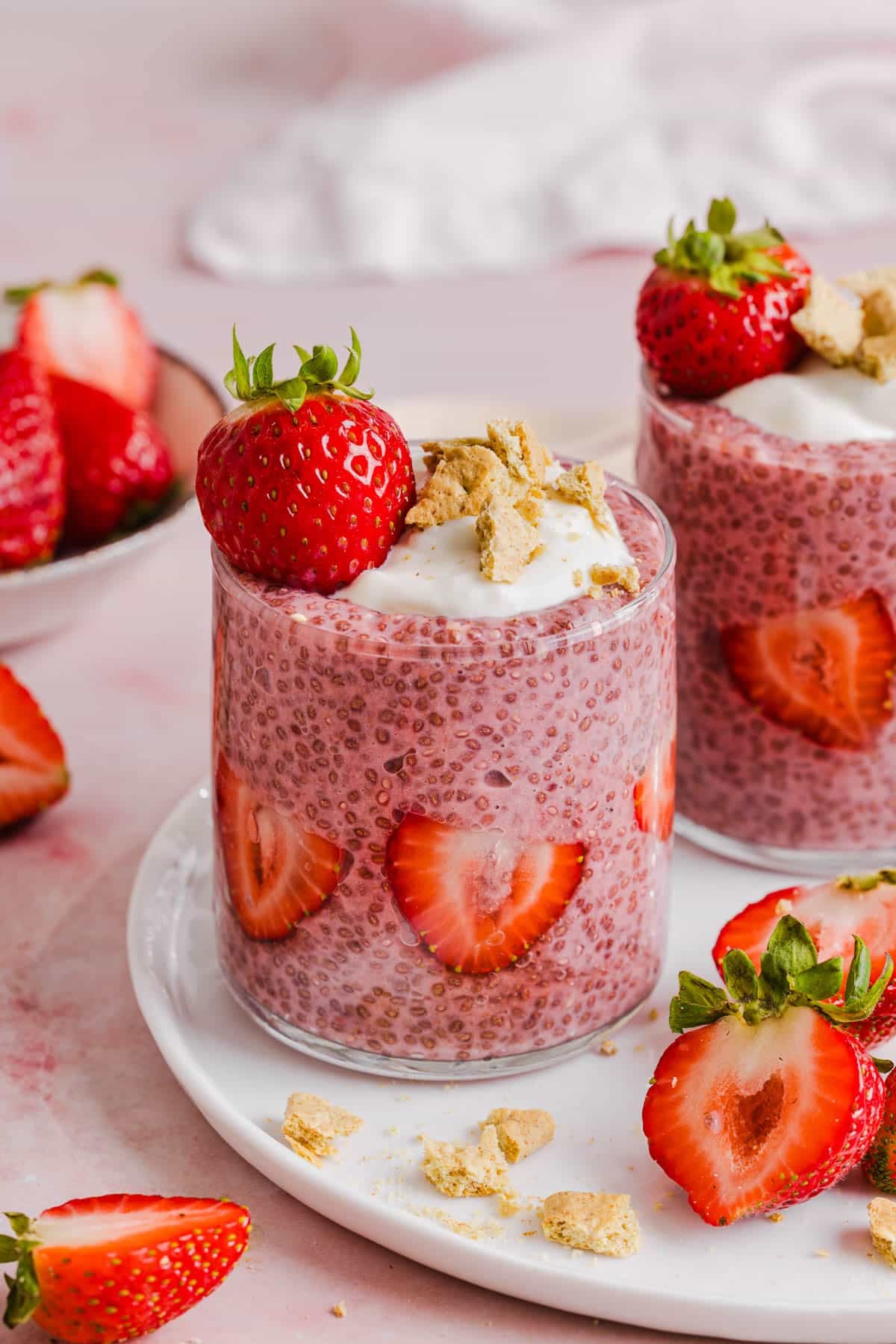 A glass of chia pudding garnished with vanilla greek yogurt, crushed graham crackers and fresh strawberries on a white plate on a countertop.