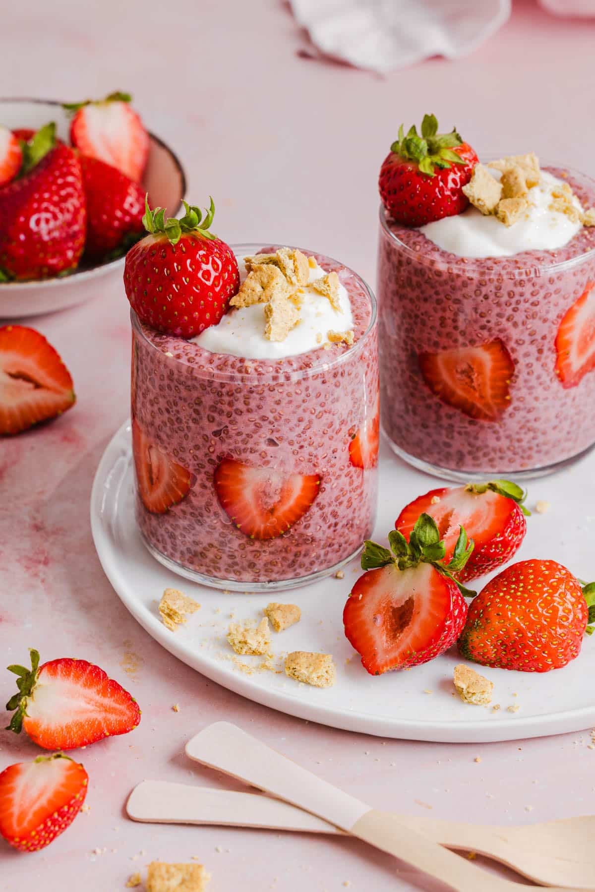 Two jars of strawberry chia pudding garnished with greek yogurt, graham cracker crumbs and fresh strawberries sitting on a white plate on a countertop.