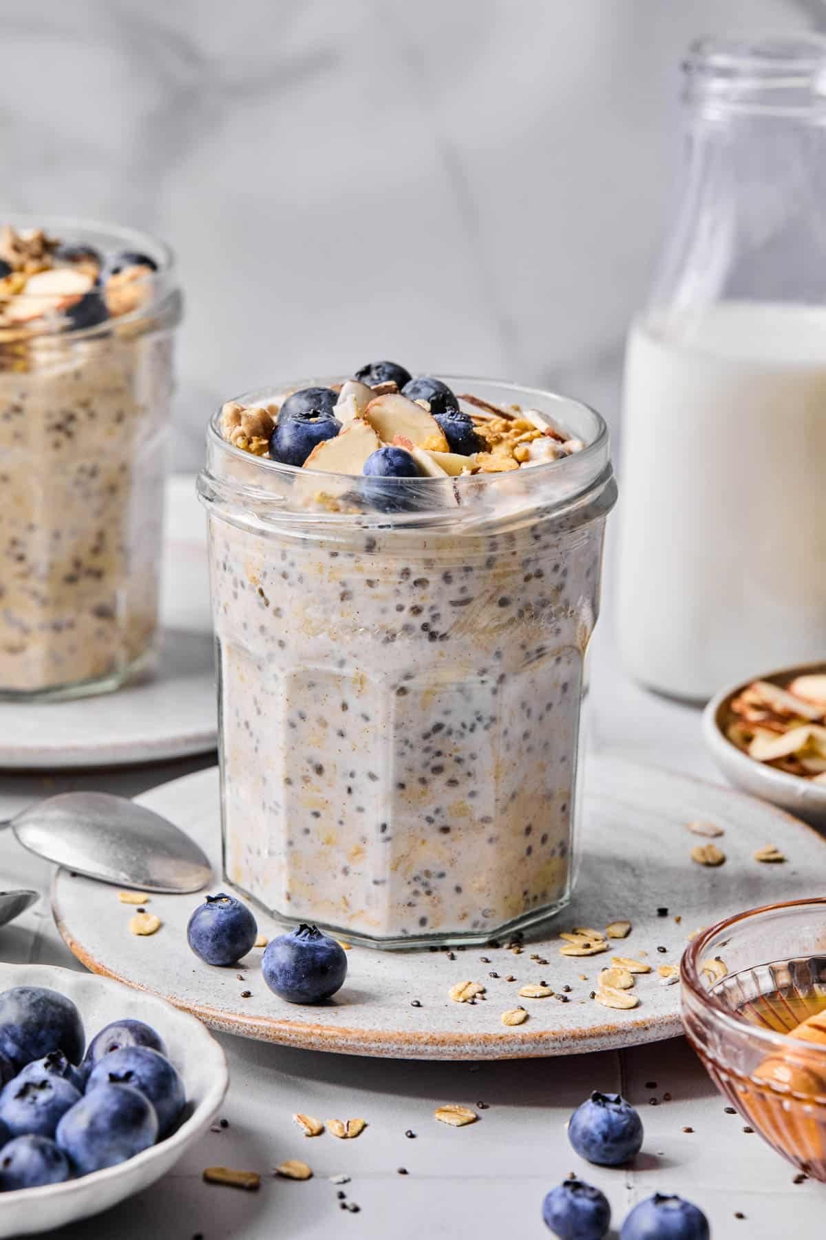 A jar of vanilla overnight oats with granola and fresh blueberries topping sitting on a serving plate on a countertop.