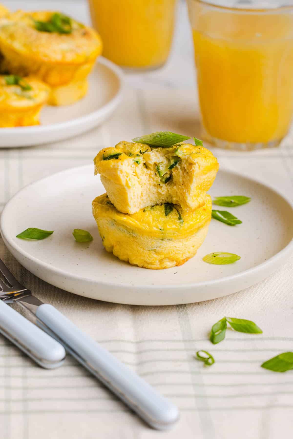 A zucchini egg muffin with a bite taken out of it sits on top of another zucchini egg muffin on a white plate on a countertop.