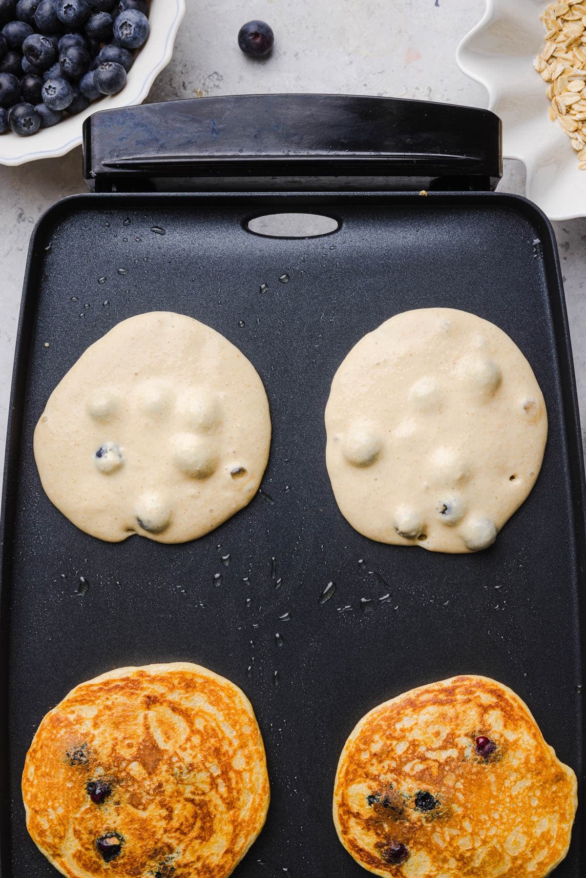 Four pancakes on a griddle for cooking, two are flipped over to finish cooking on both sides.