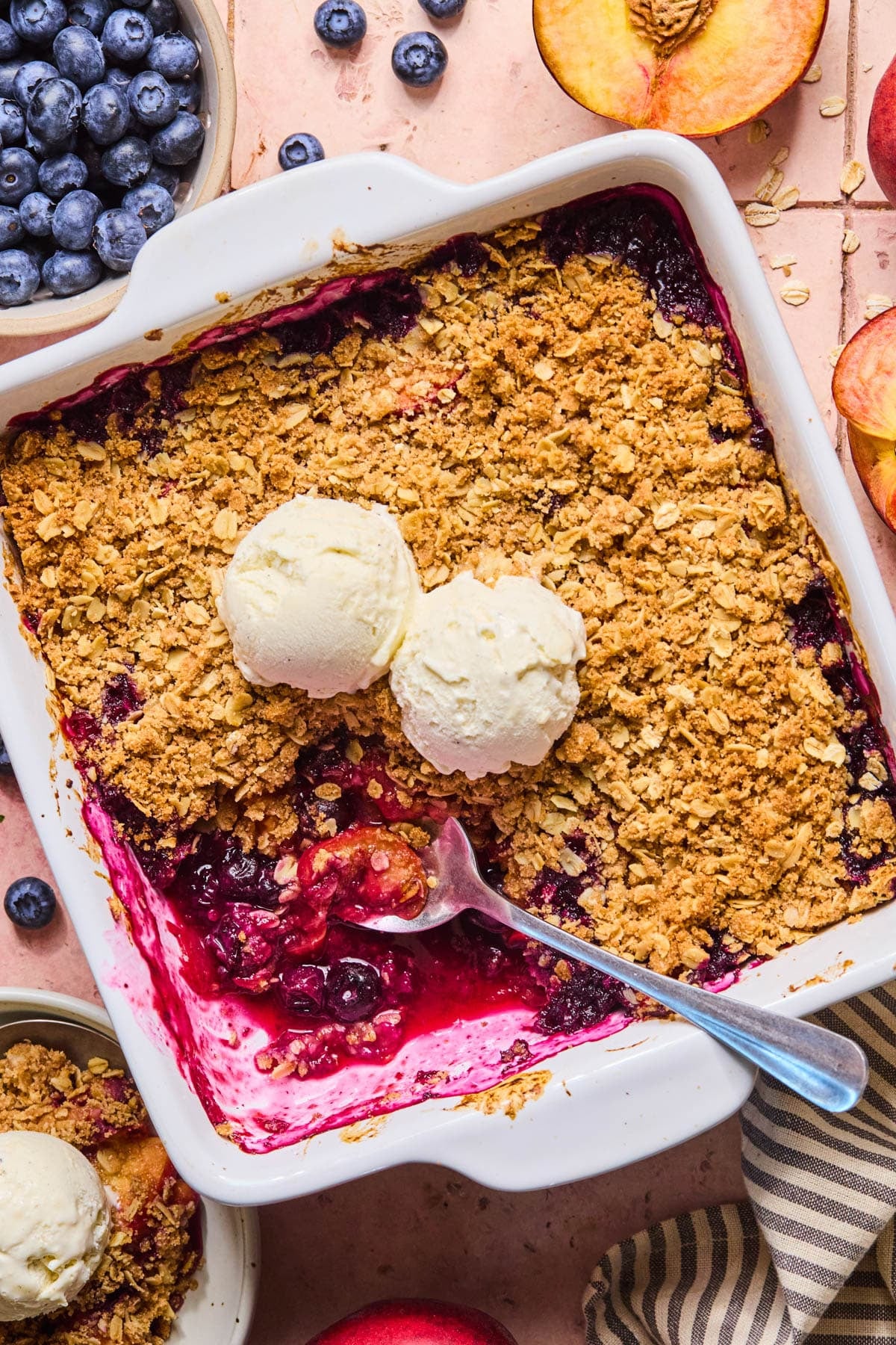 A spoon rests in the baking dish with the peach blueberry crumble with two scoops of vanilla ice cream on top.