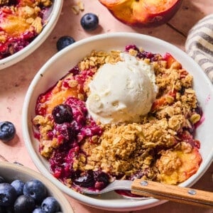 The baked peach blueberry crumble in a white bowl with a scoop of vanilla ice cream on top and a spoon resting in the bowl.