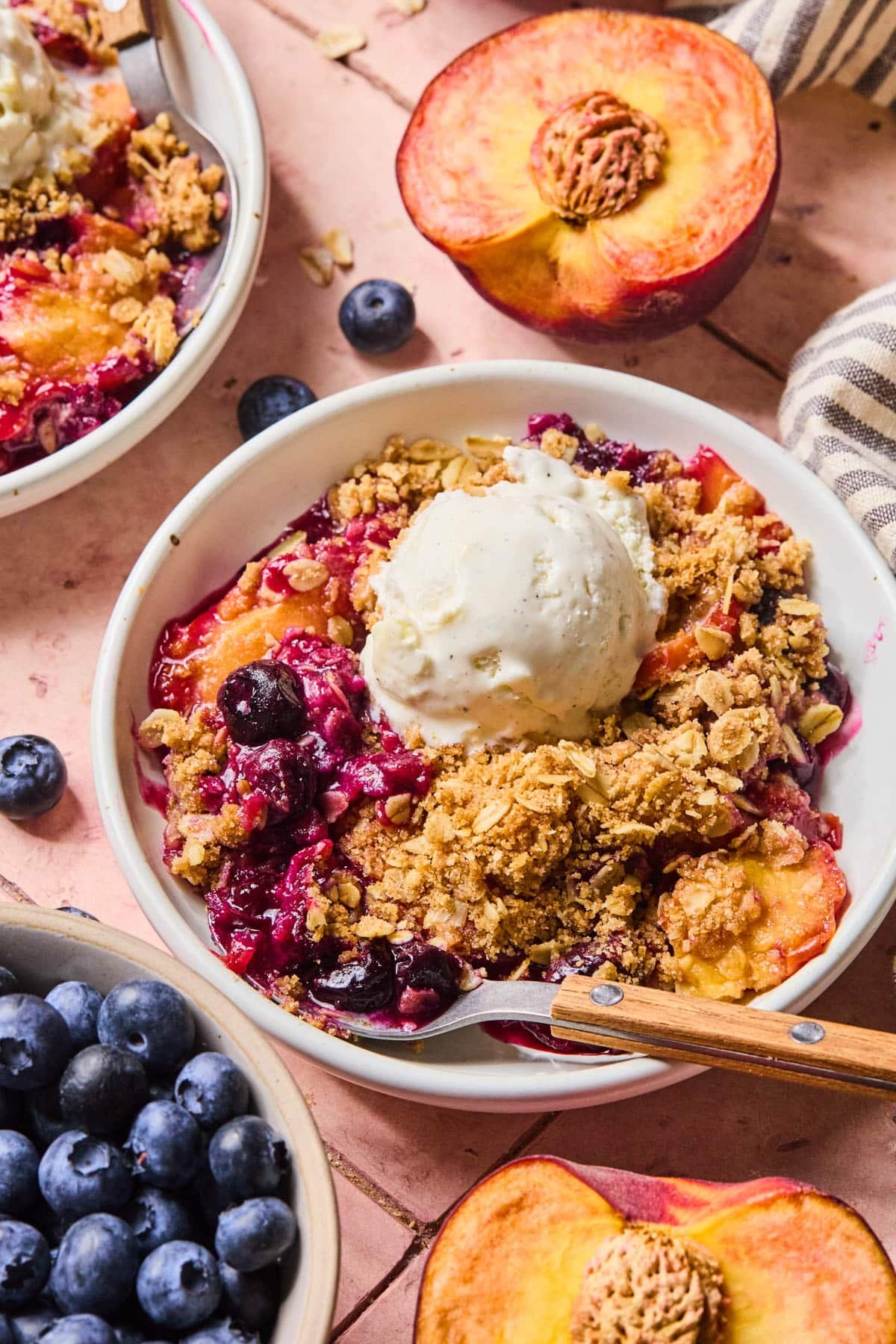 The baked peach blueberry crumble in a white bowl with a scoop of vanilla ice cream on top and a spoon resting in the bowl.