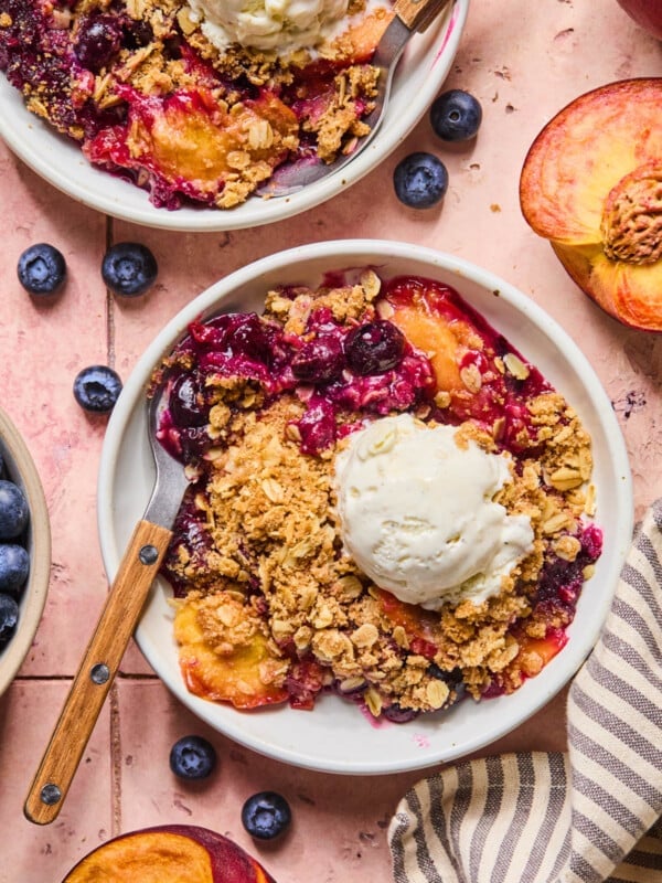 The peach blueberry crumble with a scoop of vanilla ice cream in two white bowls on a countertop.