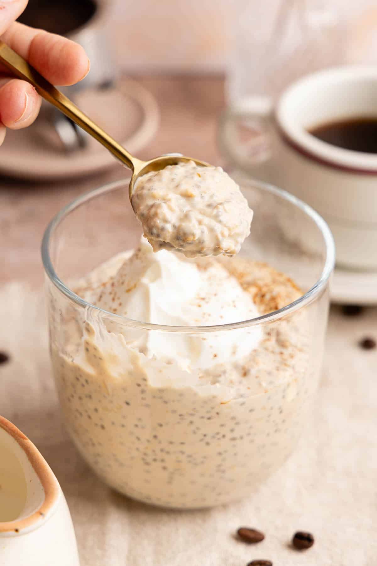 A spoon lifts coffee overnight oats from a glass sitting on a countertop.