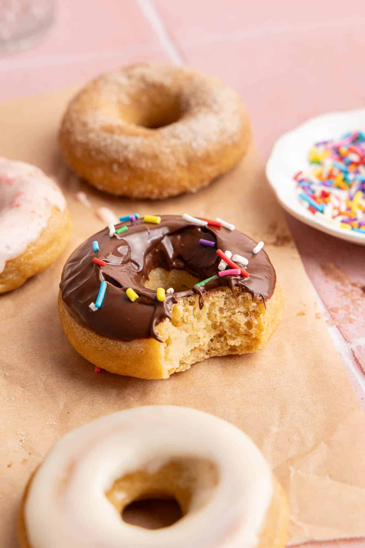 A chocolate frosted donut with a bite taken out of it sitting on a piece of parchment paper next to the other donuts.