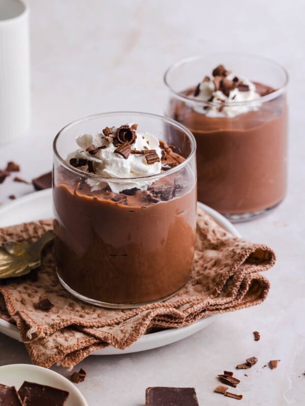 Two jars of high protein chocolate pudding on a countertop with whipped cream and chocolate shavings on top.