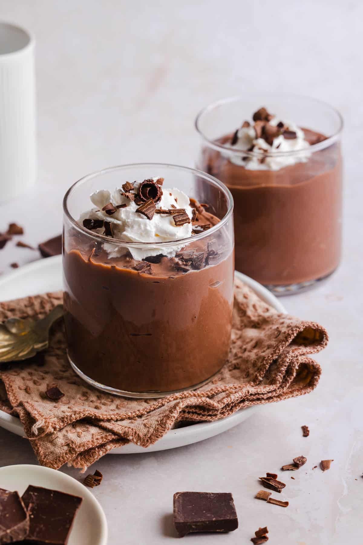 Two jars of high protein chocolate pudding on a countertop with whipped cream and chocolate shavings on top.