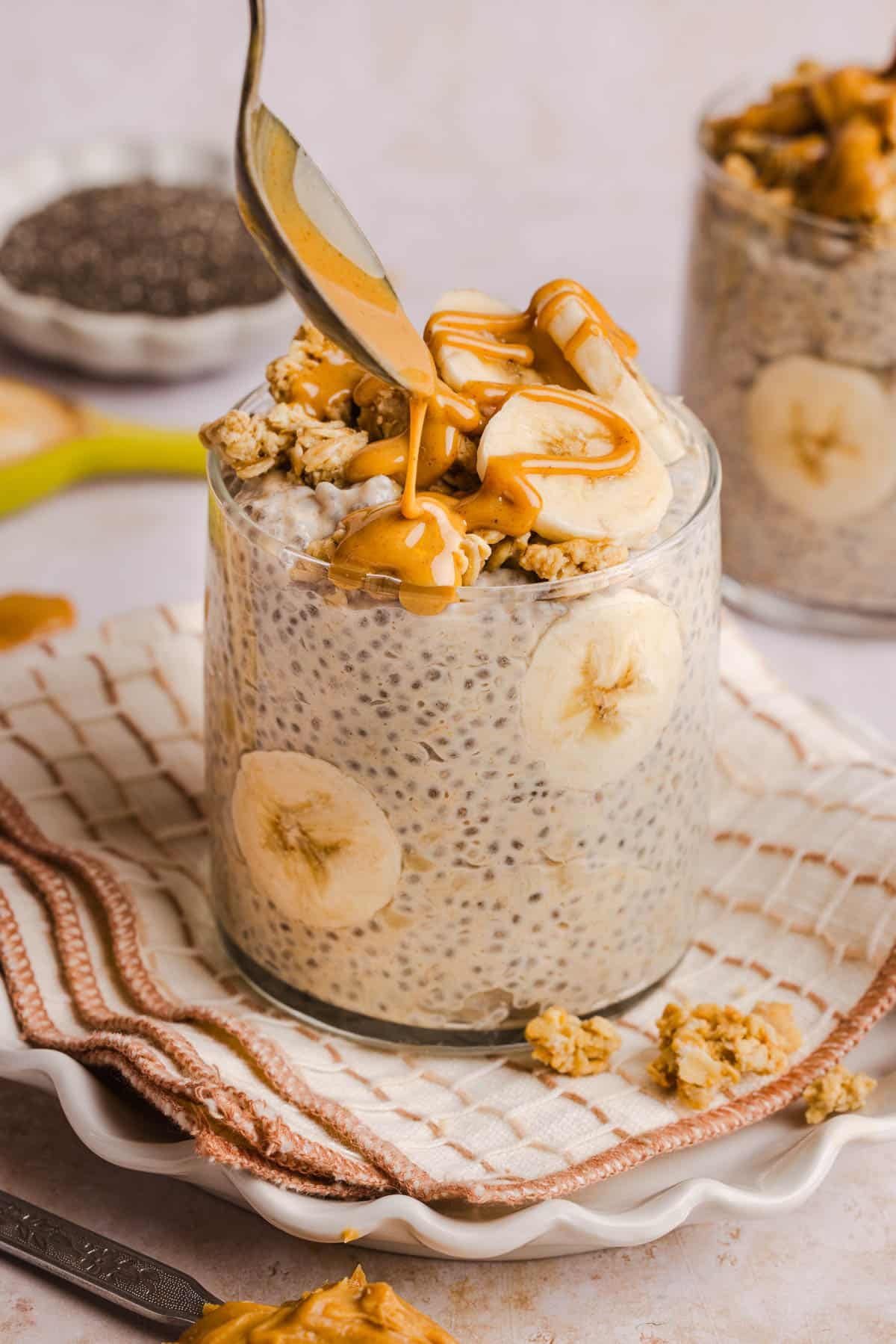 A spoon drizzles peanut butter over sliced bananas and granola on top of the chia pudding in the jar.
