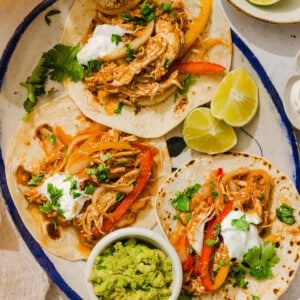 Three chicken fajitas on an oval platter with a bowl of guacamole on the side and fresh lemon wedges.