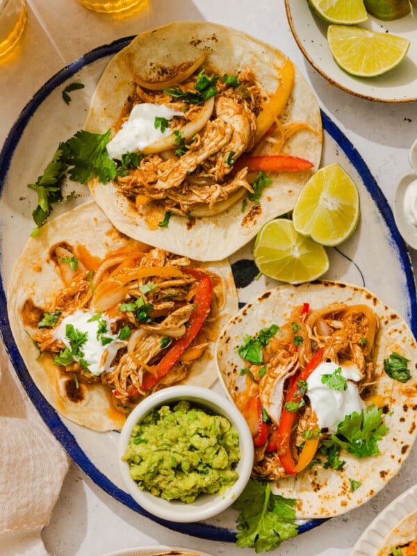 Three chicken fajitas on an oval platter with a bowl of guacamole on the side and fresh lemon wedges.