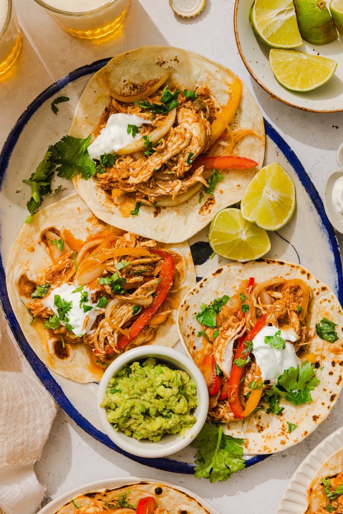 Three chicken fajitas on an oval platter with a bowl of guacamole on the side and fresh lemon wedges.