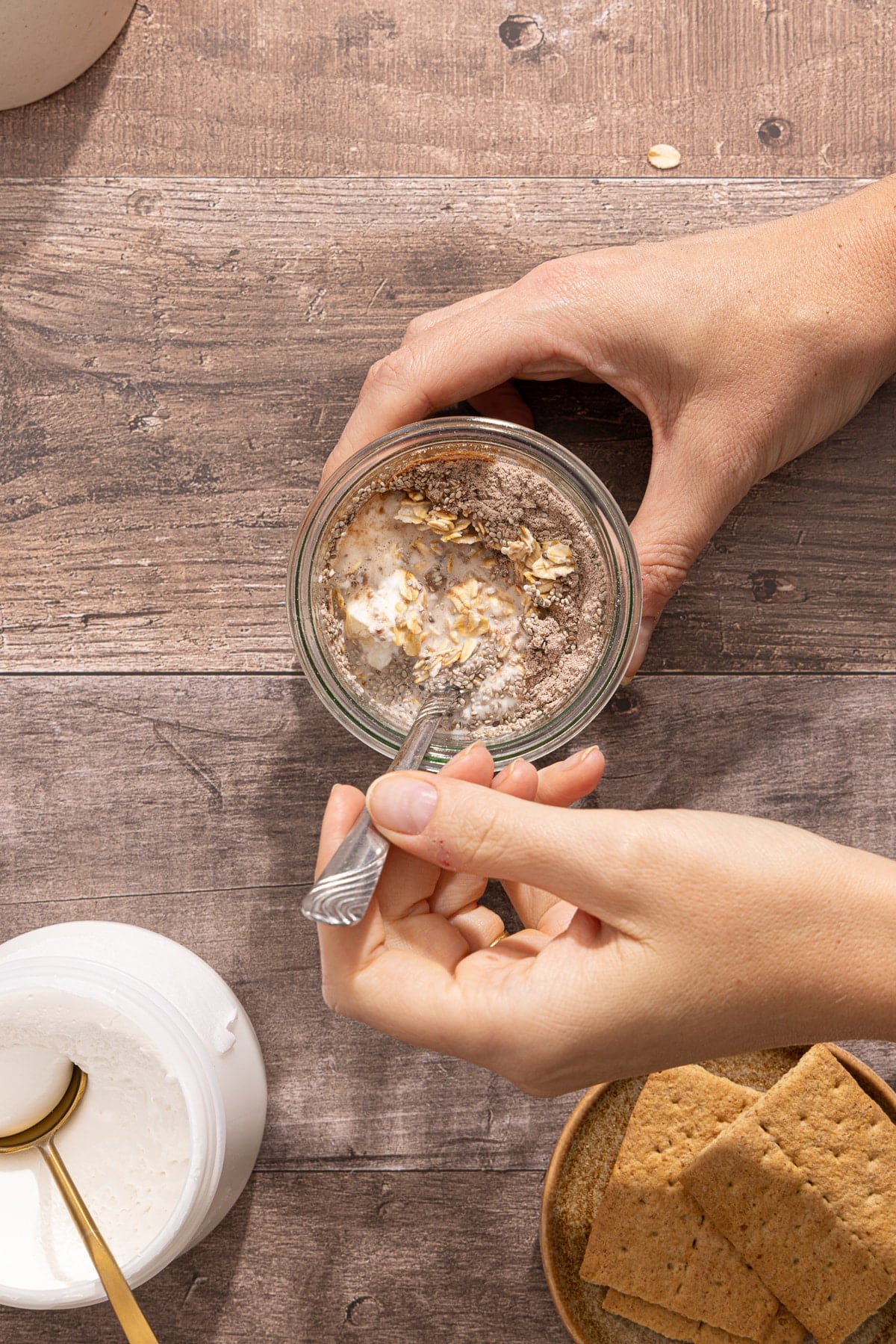 A spoon stirs the oats, milk, Greek yogurt, chia seeds, chocolate protein powder, and vanilla in a glass jar on a countertop.