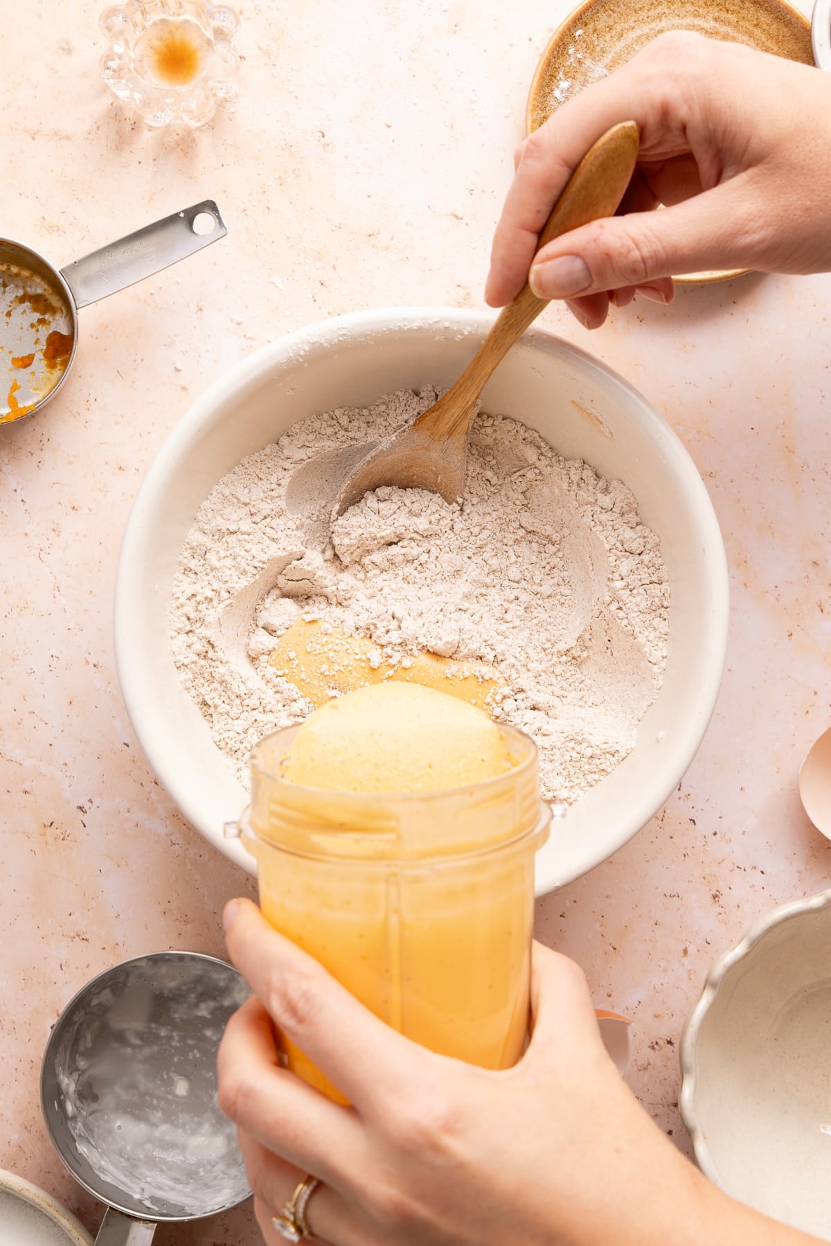 A hand pours the wet ingredients from the blender into the bowl with the mixed dry ingredients.