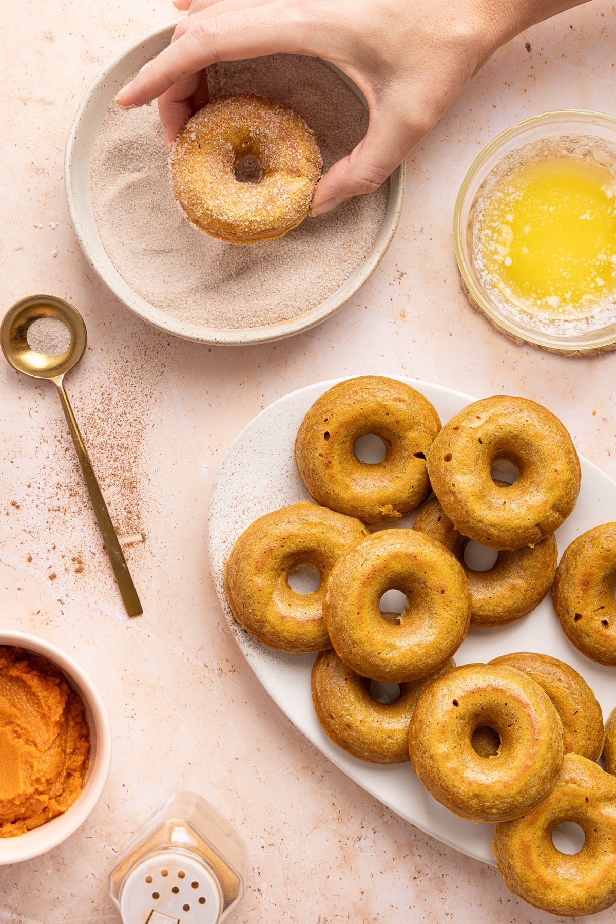 A hand dips the baked donut with butter into the cinnamon sugar mixture next to a plate of the remaining baked pumpkin donuts.