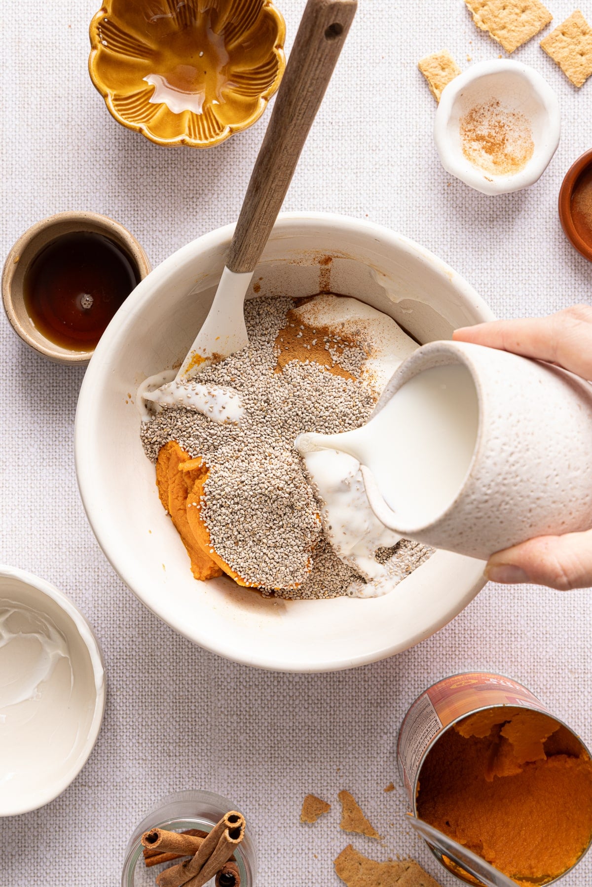 A hand pours the milk into a bowl with all of the remaining ingredients for pumpkin chia pudding.